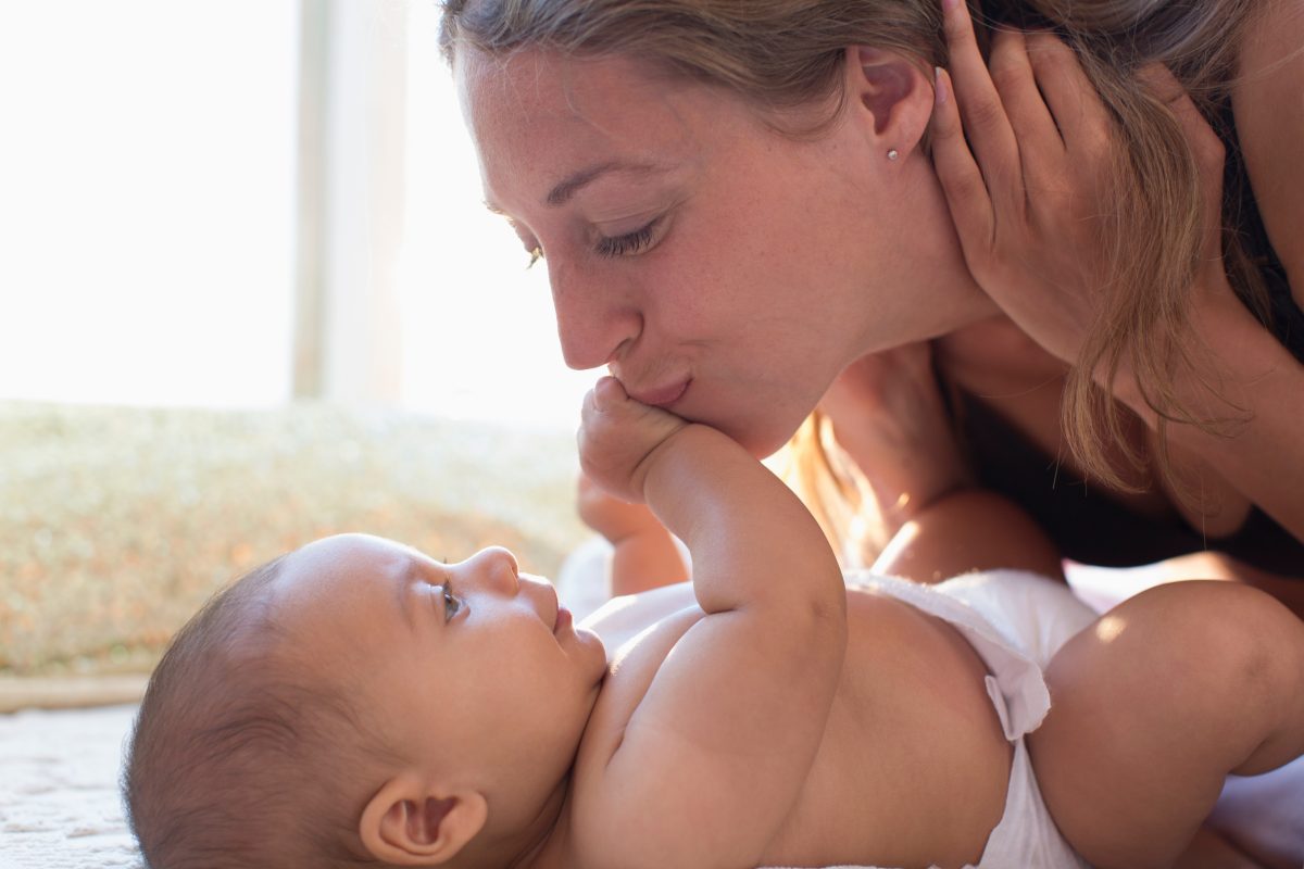 Baby ligger på rygg. Mamma lutar sig fram så att babyn kan ta på mammas mun. Mamma tittar på babyns ansikte. Kärlek.